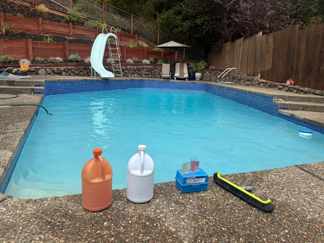 Pool chemicals test kit and brush on deck beside clean blue pool with slide ready for weekly maintenance service