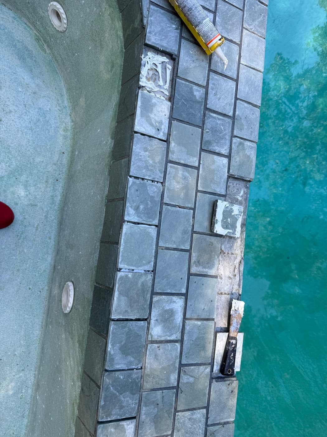 Pool coping tile repair in progress with adhesive and replacement stone tiles being set between pool and spa